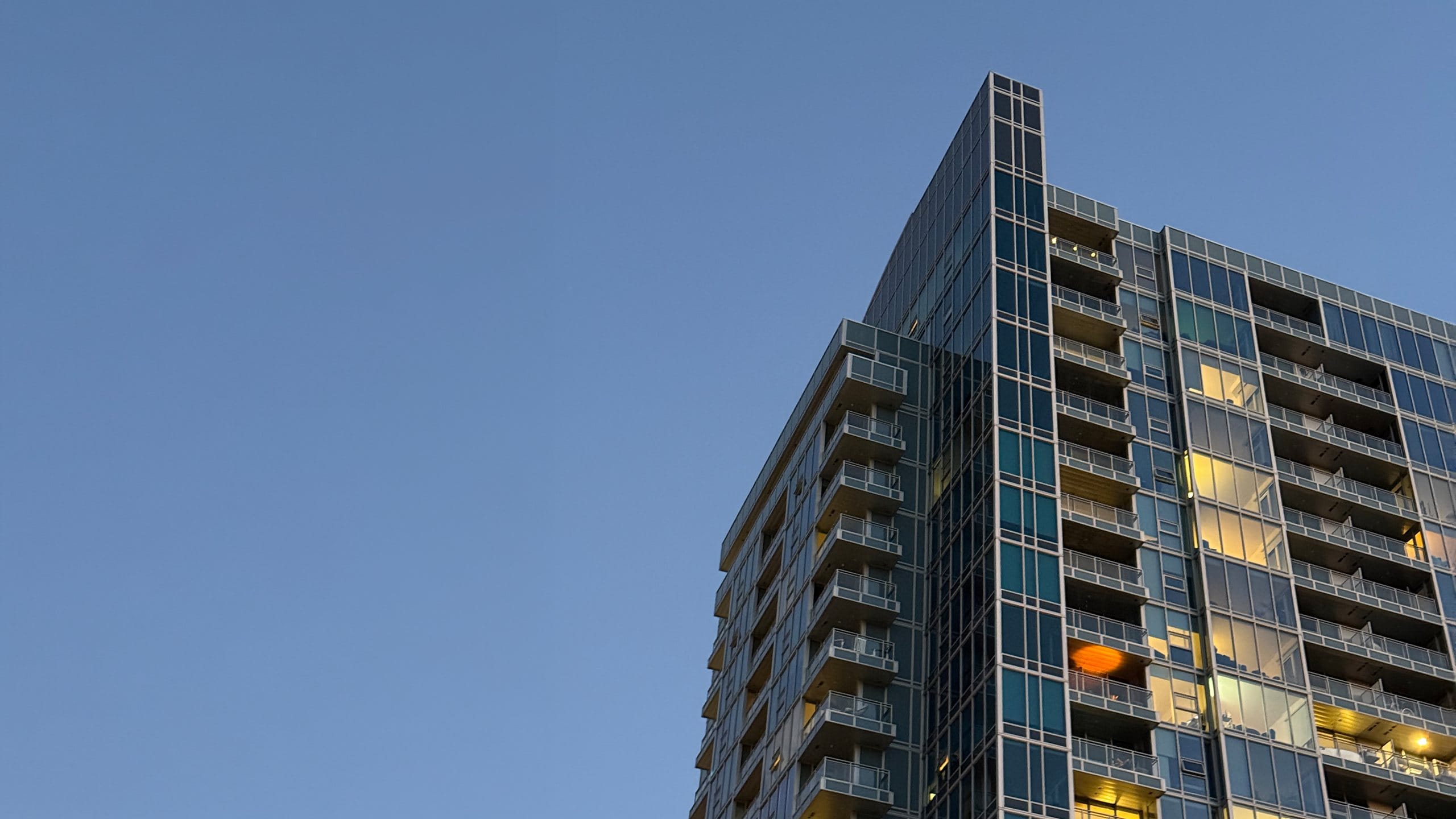 A glass residential tower photographed from street level at blue hour, its lit corner rising into a clear sky